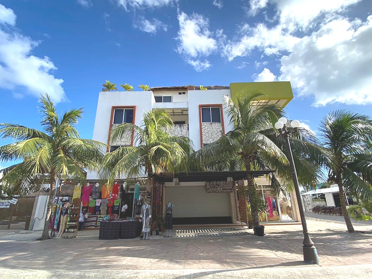 A three-story building is seen under a clear blue sky, framed by palm trees. The front showcases a shuttered store display, with colorful items visible. The structure features a modern design, with bright green accents and a balcony on the upper level.