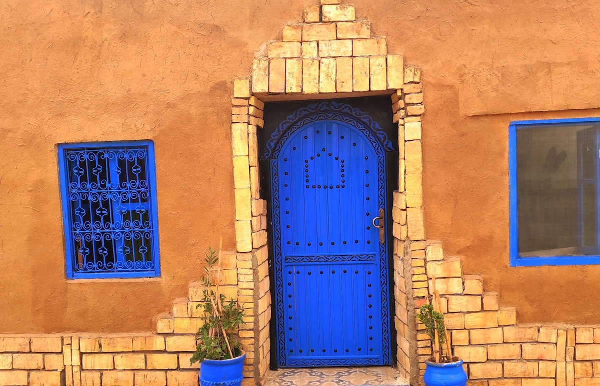 A traditional adobe structure features a striking blue door framed by intricately designed stonework. The entrance is complemented by blue pots containing greenery on either side. A decorative window with metal grilles enhances the facade, showcasing local architectural style and craftsmanship.