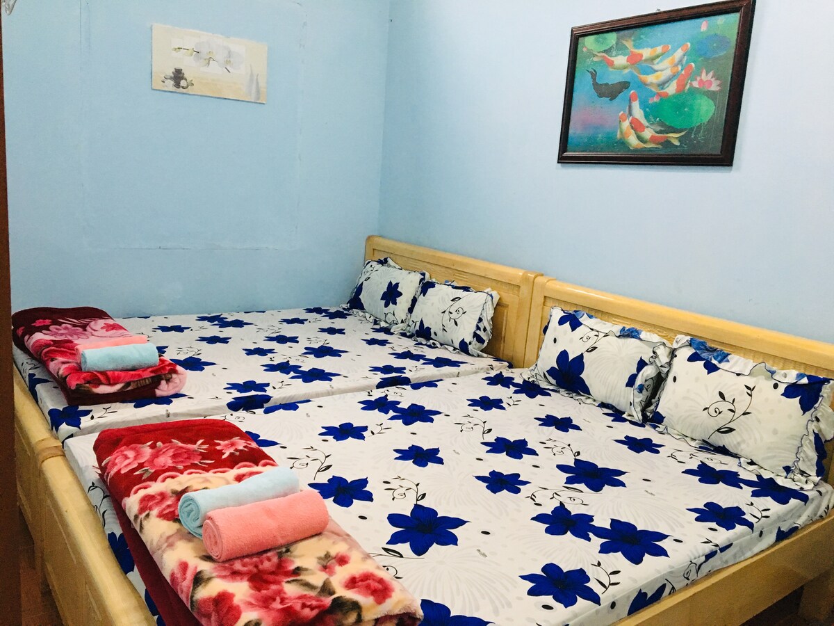The sleeping area features two double beds with floral-patterned bedding and decorative pillows. Soft towels are neatly arranged on the beds. A colorful painting adorns the wall, adding a touch of vibrancy to the serene blue room.