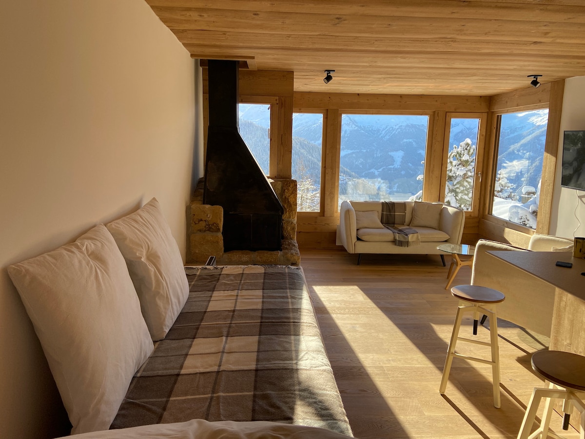 A spacious living area features large windows offering panoramic views of the mountains. A cozy fireplace is positioned next to a lounging area with a white sofa. A dining table and light chairs are placed nearby, complemented by warm wood accents throughout.