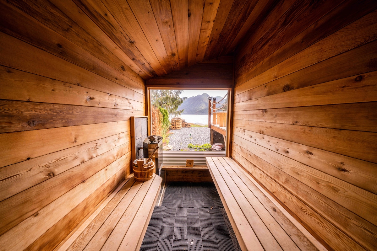 A wooden sauna room features natural wood-paneled walls and benches, with large windows providing views of the ocean and mountains. The interior includes a wooden bucket and ladle, reflecting a serene atmosphere for relaxation.