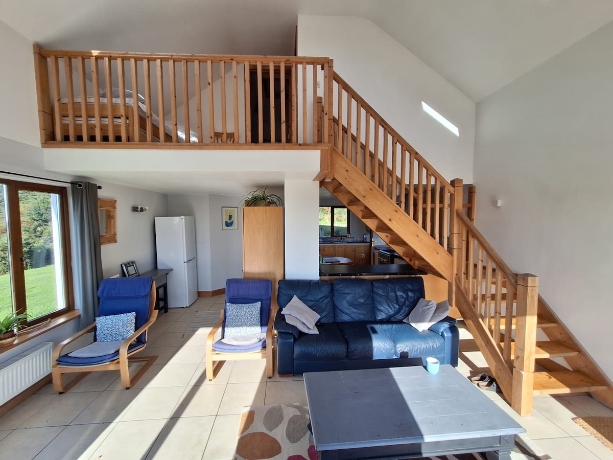 An open living area features a blue sofa and two lounge chairs, with a staircase leading to an upper gallery space. Large windows allow natural light to flood the room, highlighting the kitchen and dining area in the background.