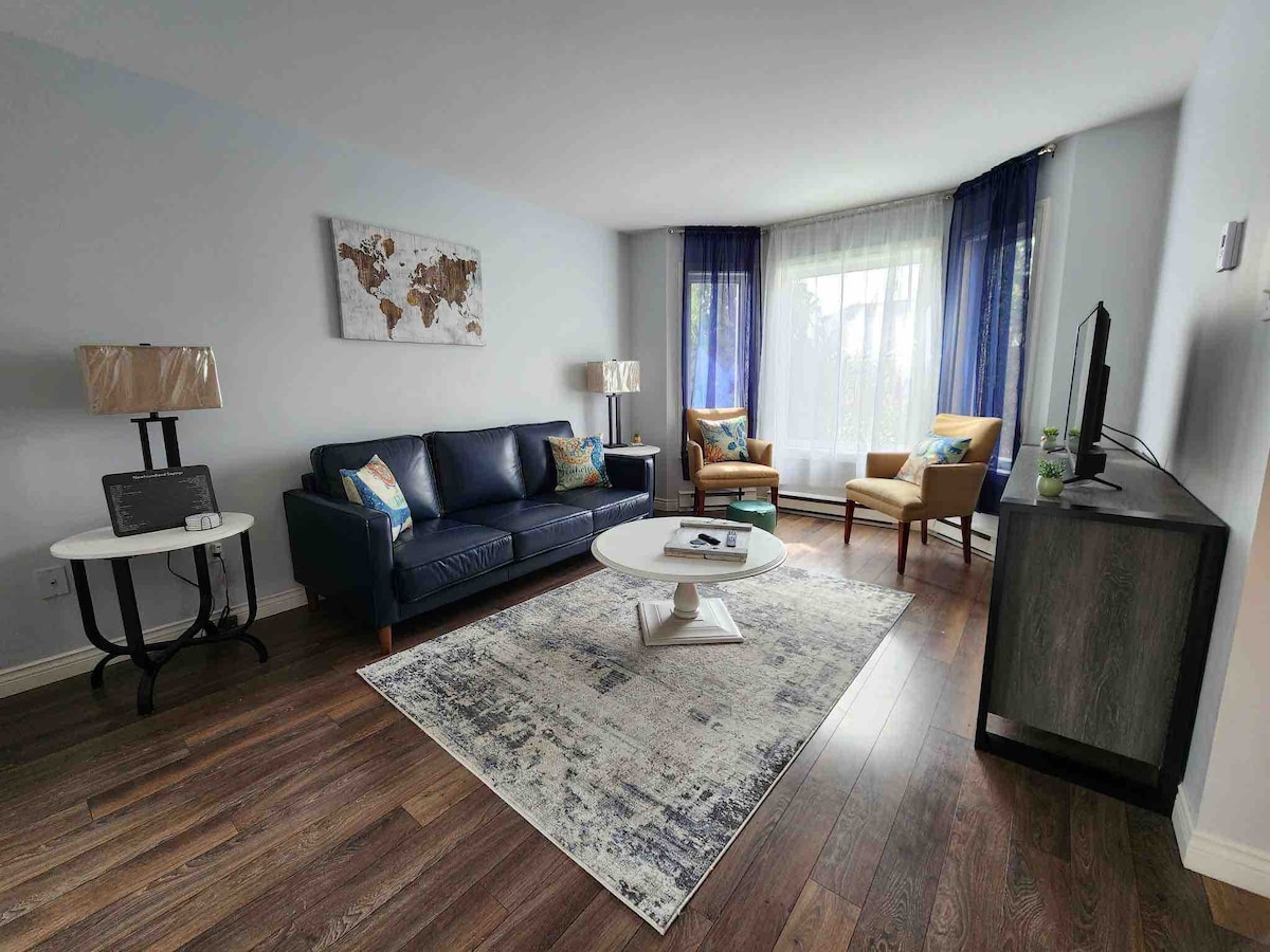 A living room features a comfortable navy blue sofa paired with two accent chairs. A round coffee table sits on a textured area rug. Natural light filters through sheer curtains, illuminating a wall with a map motif and a television stand positioned nearby.