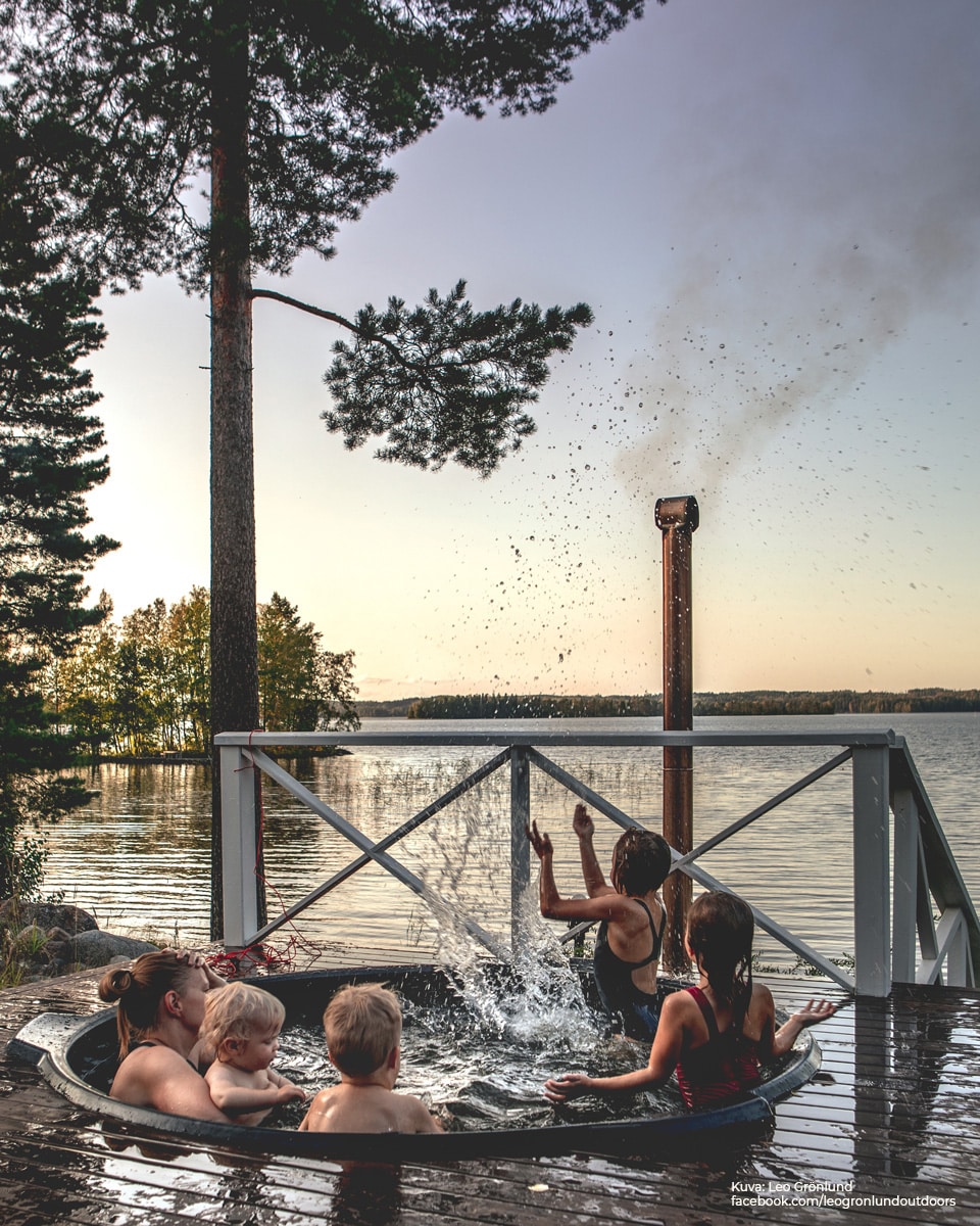 A six-person hot tub is set on a wooden deck overlooking the lake, surrounded by trees. Water splashes in the evening light, creating a lively atmosphere as individuals enjoy their time together. A sauna chimney is visible in the background, adding to the serene lakeside setting.