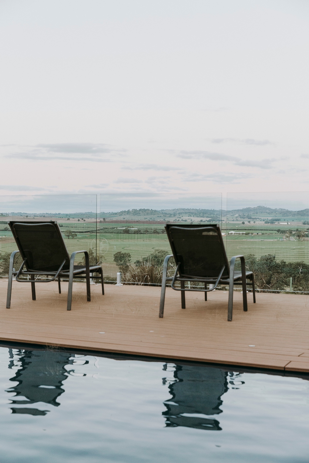 Two lounge chairs are positioned beside a sleek pool. The chairs are oriented towards expansive views of the countryside, with rolling hills and a soft horizon in the background. The pool's surface reflects the chairs and the sky above.