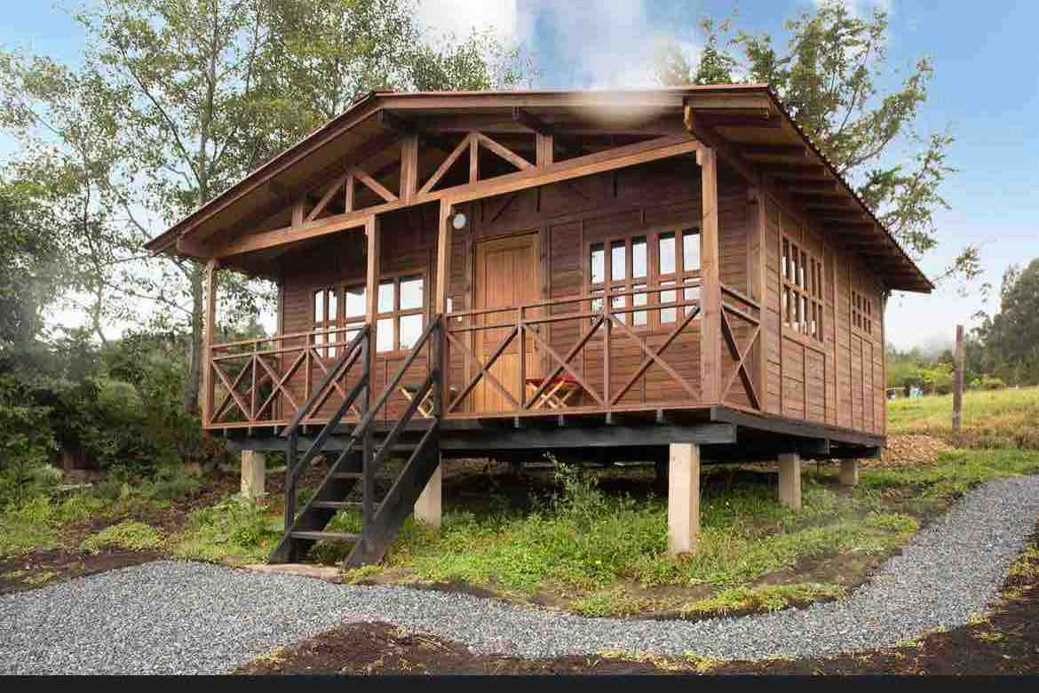 A wooden cabin is shown, elevated on pillars with stairs leading to the entrance. Large windows provide natural light, framed by wooden beams. Surrounding greenery enhances the tranquil setting, with a gravel pathway leading towards the cabin.