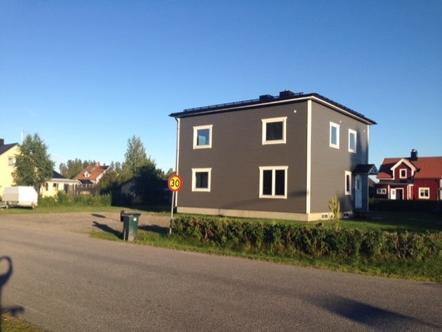 A modern two-story house is shown from the street view, featuring a grey exterior and multiple windows. The surrounding area includes greenery and residential homes, with a clear blue sky above. A recycling bin is positioned in the foreground.