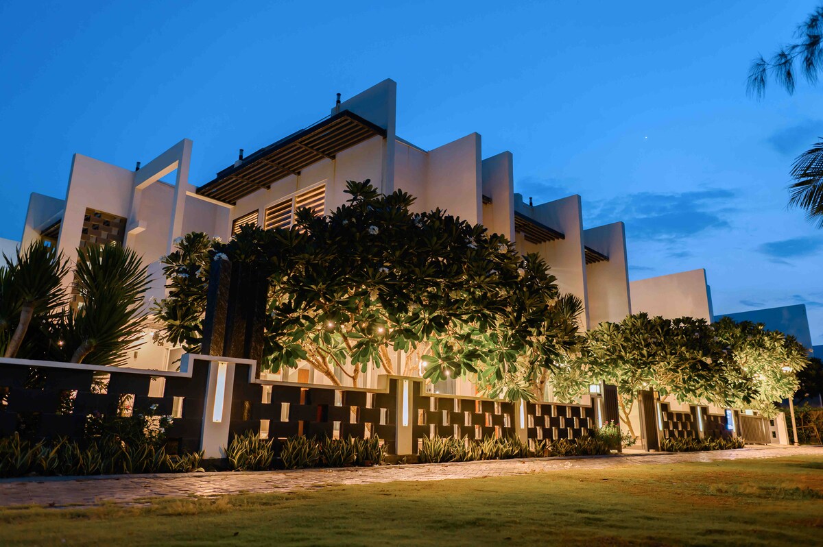 The exterior of a modern villa is depicted at twilight, showcasing clean lines and a sleek design. Soft lighting highlights the lush greenery and landscape, creating a serene atmosphere. The surrounding environment is well-maintained, with a pathway guiding the viewer’s eye towards the entrance.