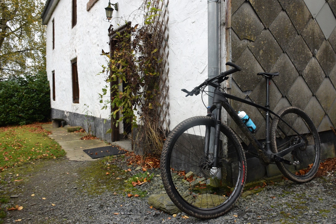 A bicycle is positioned next to the exterior wall of a historic house, partially covered in climbing vines. The entrance is framed by a walkway and bordered with gravel, while autumn leaves are scattered across the ground.
