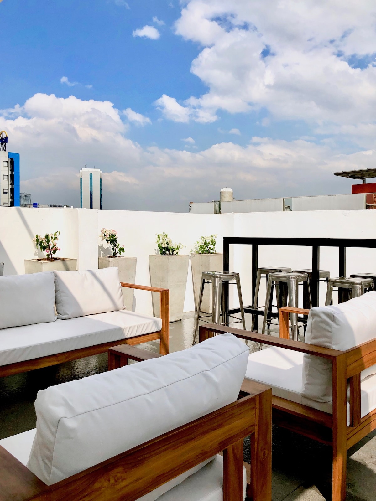 An outdoor seating area features wooden chairs with white cushions arranged around low tables. Potted plants add greenery to the space, and a row of high stools stands against a backdrop of a clear blue sky and cityscape.