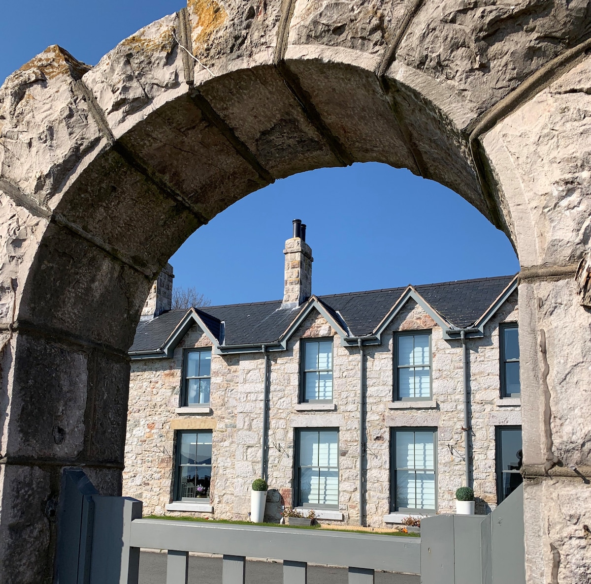 A stone archway frames a view of a charming townhouse featuring a light stone facade. Large windows are adorned with planters, and a clear blue sky serves as a backdrop. The structure displays a traditional design with a pitched roof and multiple chimneys.