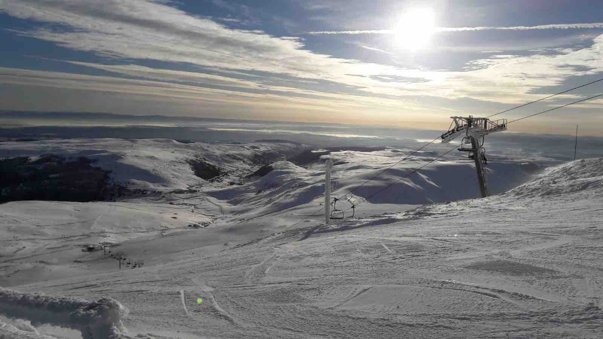 A vast snowy landscape is captured under a bright sun. Ski lifts are visible against the backdrop of snow-covered mountains, with gentle slopes and a serene sky stretching far into the distance. The scene conveys a peaceful winter atmosphere.