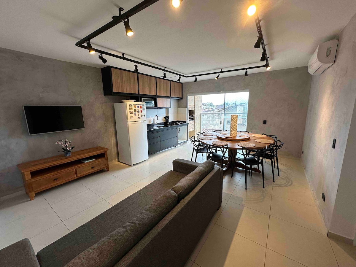 An open-concept living area features a sleek kitchen with modern appliances and a round dining table that seats eight. A comfortable sofa sits across from a wall-mounted TV, with natural light flowing in through a large window highlighting the neutral color palette.