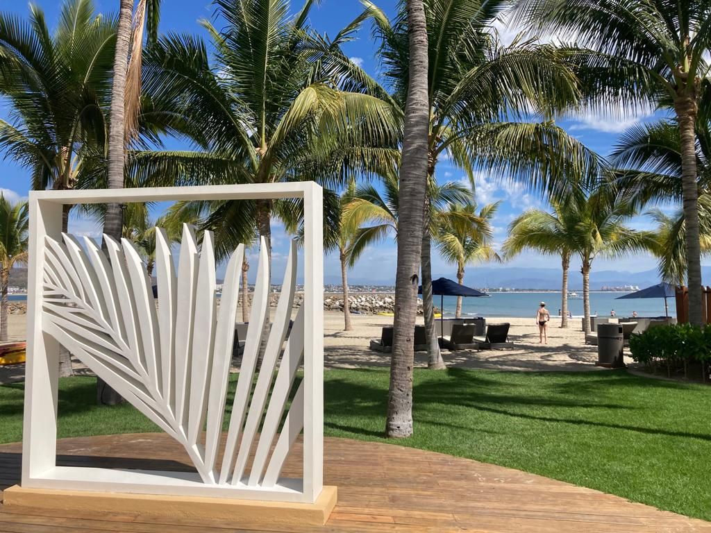 A decorative white sculpture shaped like palm leaves is positioned on a wooden deck. Lush green grass surrounds the area, and tall palm trees frame the view of the sandy beach and calm ocean beyond. Individuals can be seen enjoying the beach in the background.