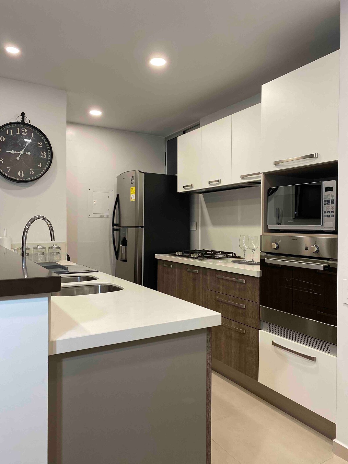A modern kitchen is equipped with stainless steel appliances, including a refrigerator and microwave. Cabinets in a combination of light and dark finishes provide ample storage. A large clock adorns the wall, while ambient lighting enhances the workspace.