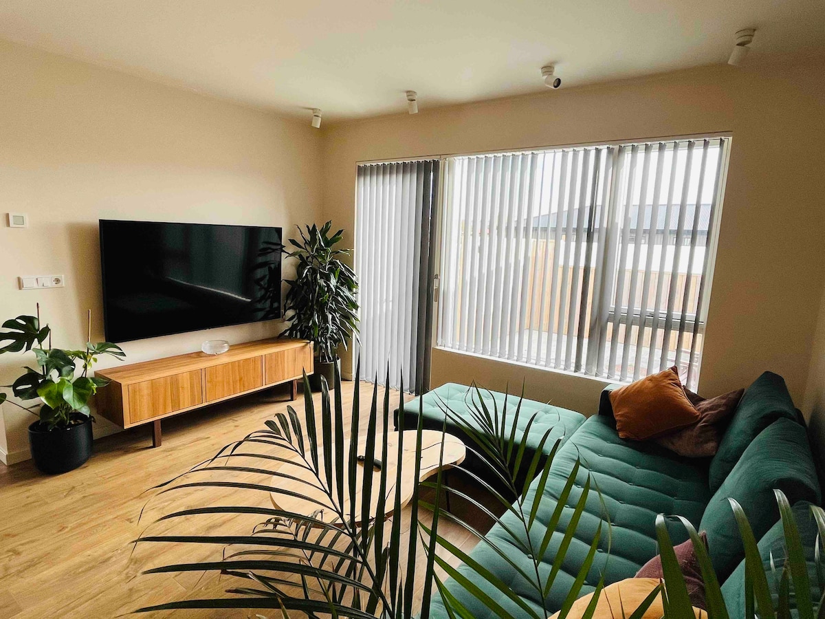 A spacious living room features a large television mounted on a light-colored wall, with a wooden media console below. A sectional sofa in a rich green hue is accented by decorative pillows. Natural light filters through vertical blinds, illuminating indoor plants positioned nearby.