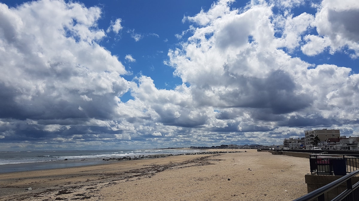 A wide view of a sandy beach stretches under a sky filled with large, fluffy clouds. The ocean is seen gently lapping at the shore, while distant buildings are visible along the coastline. The boardwalk runs parallel to the beach, offering accessibility to the area.