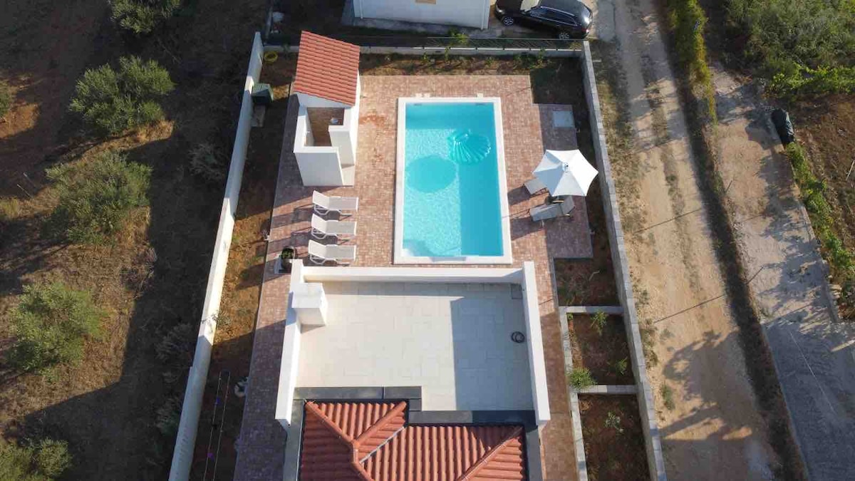 An aerial view of the property reveals a private swimming pool surrounded by a spacious terrace. White lounge chairs are arranged by the edge of the pool, while a shaded seating area is visible nearby. The well-maintained grounds are framed by greenery and pathways, providing access.