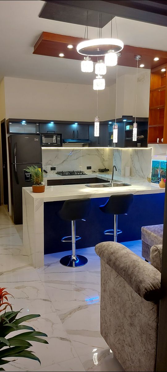 A modern kitchen area is visible, featuring a marble countertop with two high-backed stools. The cabinetry is dark, illuminated by soft lights above. A built-in stove and refrigerator are present, with decorative plants adding a touch of color to the space.