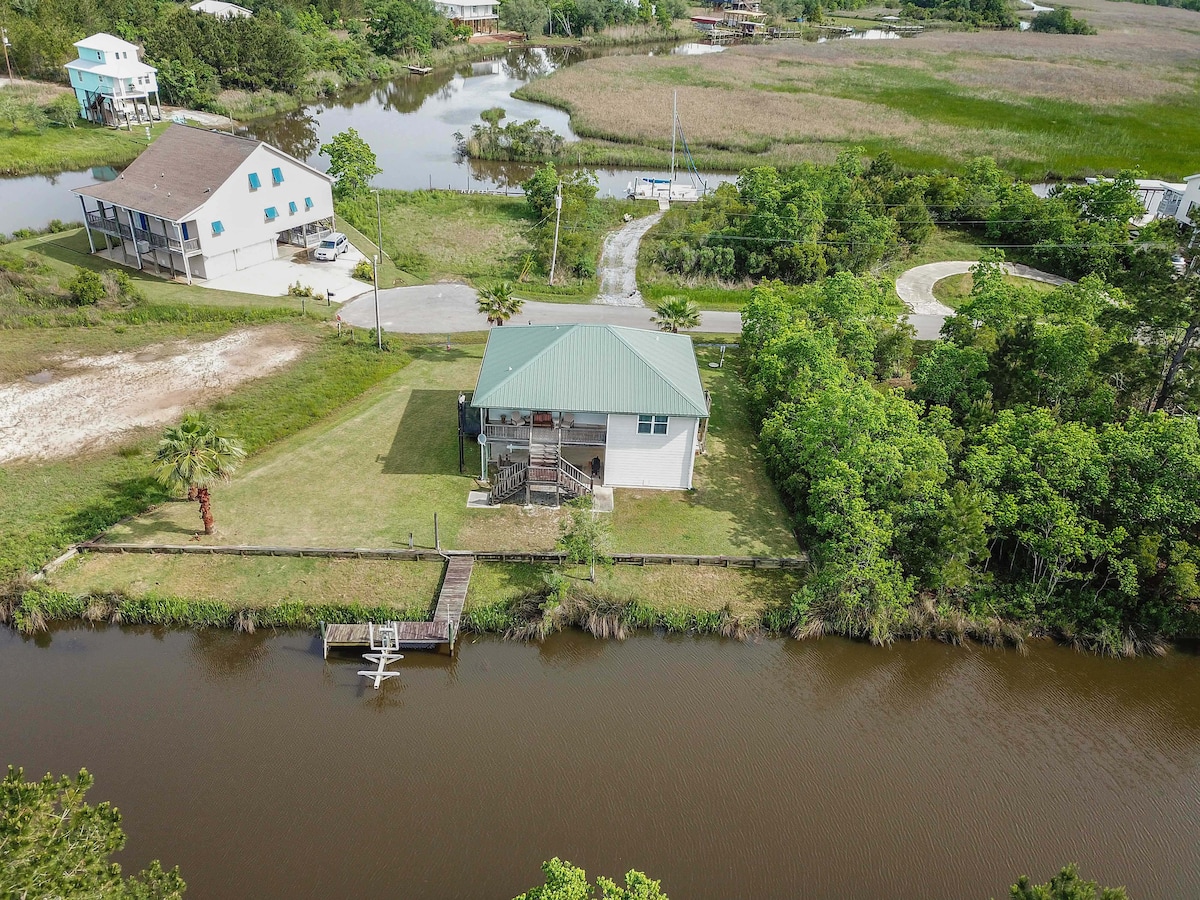 An aerial view of Lazy River House showcases its positioning on the canal's banks, surrounded by lush greenery. The property features a dock for boating, while the nearby water reflects the natural landscape and the home’s distinct green roof.