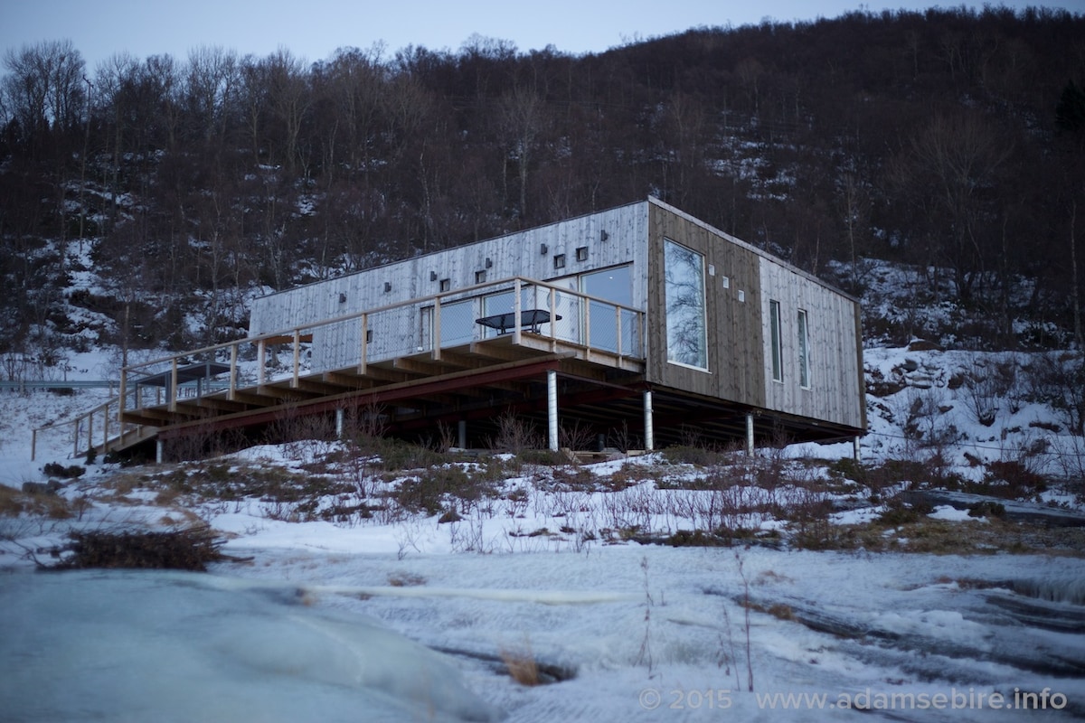 A wooden cabin is elevated on stilts, surrounded by a snow-covered landscape. Large windows reflect the surrounding nature, while a wooden deck offers an outdoor space. The structure has a modern design, with clean lines and a natural wood finish.