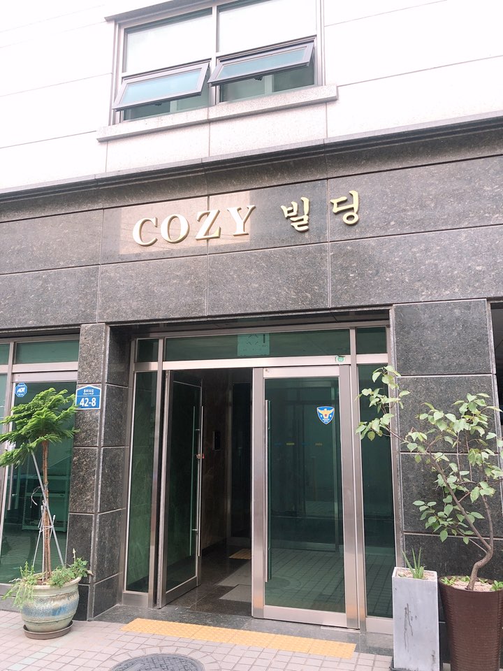 The entrance of the building is marked with a gold sign that reads 'COZY' on a dark stone facade. Large glass doors provide access, flanked by small trees in planters, creating a welcoming appearance to the entryway.