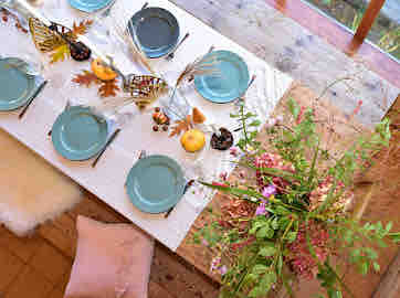 A large dining table is set with blue plates and silver utensils, surrounded by eight chairs. A centerpiece of flowers and natural elements enhances the setting. Soft textures from cushions and a fur throw are visible, contributing to a warm atmosphere.