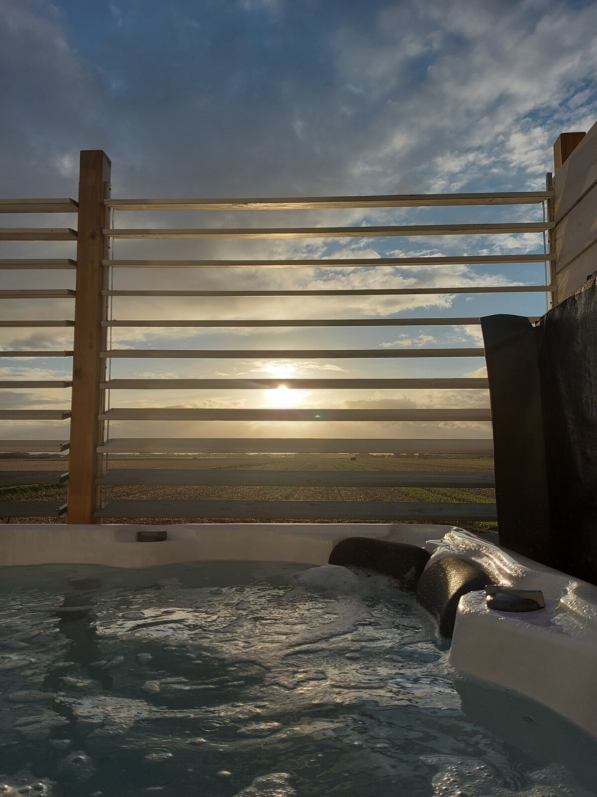 A jacuzzi is visible, surrounded by wooden slats that provide partial privacy. A serene sunset casts soft light over the landscape, with rolling fields seen in the background. The bubbling water glistens, creating a relaxing ambiance.