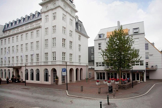An urban scene showcases two buildings with distinctive architectural styles. The left structure features arched windows and a decorative cornice, while the right building displays a modern facade with large windows and a terrace. A parking area is visible, along with greenery adding to the setting.