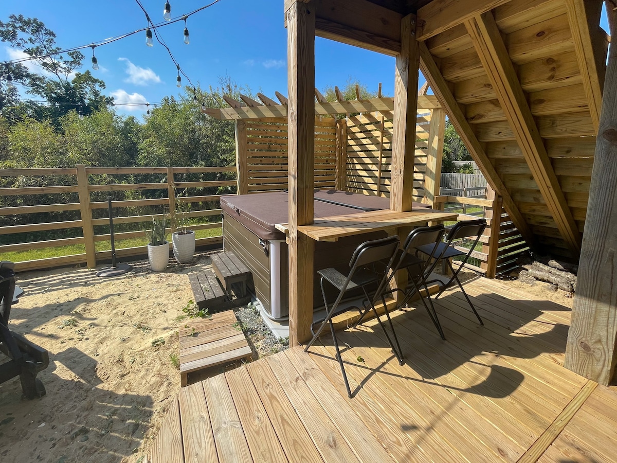 An inviting outdoor space features a hot tub situated on a wooden deck, surrounded by greenery. Two chairs are positioned near a wooden table, offering a spot to relax. Soft lighting is provided by hanging string lights overhead, enhancing the outdoor atmosphere.