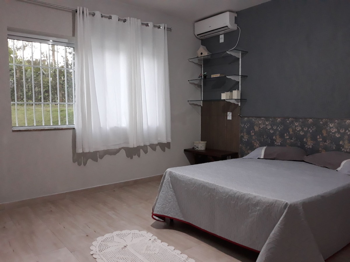 A bedroom is presented with a spacious bed featuring soft grey bedding. Light-colored curtains filter natural light through a window, while a minimalistic shelf on the wall holds decorative items. A small table is positioned beside the bed, contributing to the room's clean aesthetic.