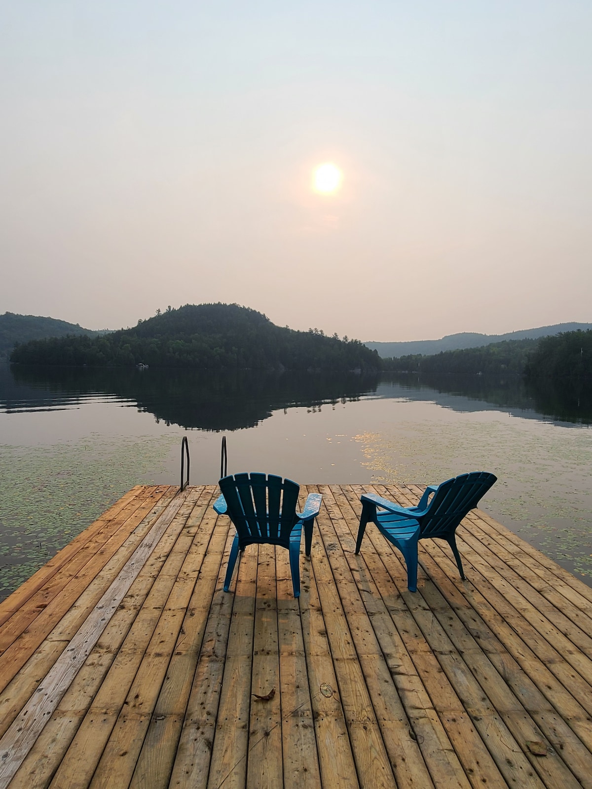 A wooden dock extends over calm water, featuring two blue adirondack chairs facing the horizon. The sun hangs low in the sky, casting a soft glow over the tranquil landscape, while hills outline the background in gentle silhouettes.