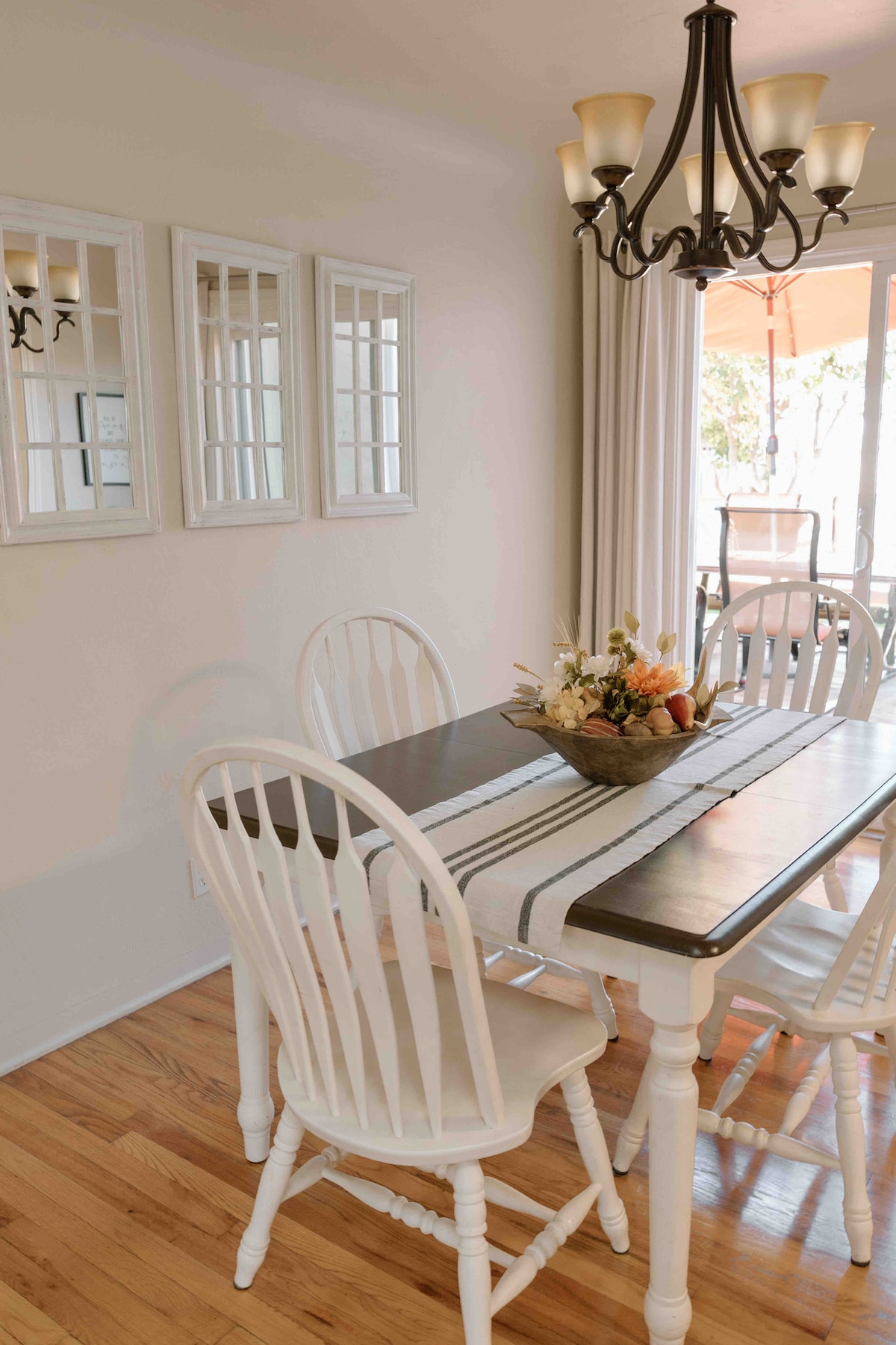 The dining area features a wooden table surrounded by white chairs. A centerpiece with flowers and fruit is present on the table. Three framed mirrors hang on the wall, and a chandelier provides overhead lighting. A sliding door leads to an outdoor patio.