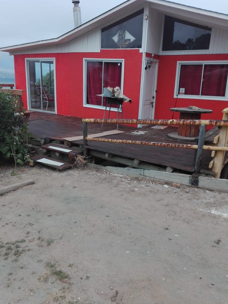 A vibrant orange house is accompanied by a spacious wooden deck, featuring a small seating area and a table. Large windows allow natural light to illuminate the interior, while a path leads from the sandy area to the entrance of the home.