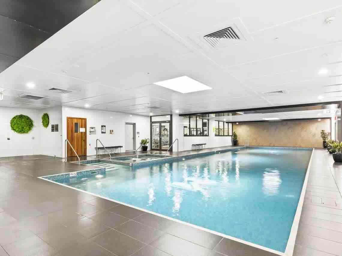 The indoor swimming pool features a large, clear blue water surface surrounded by a tiled deck. Natural light filters in from overhead skylights, and potted plants add a touch of greenery. Relaxation areas are included along the edges for guest comfort.