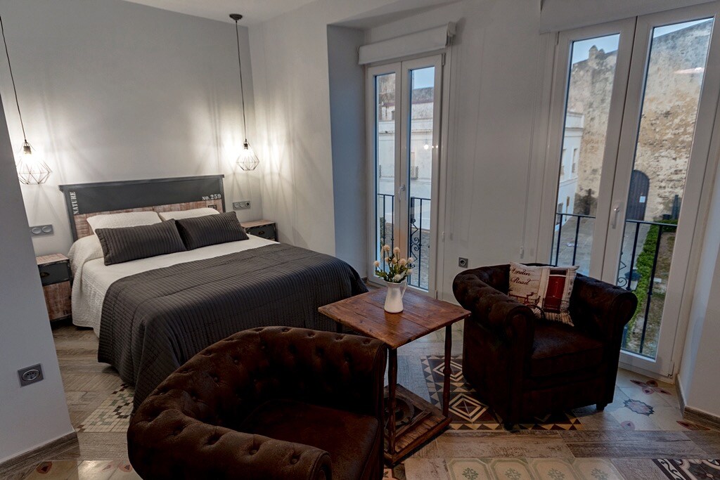 A well-appointed bedroom features a spacious bed with neutral-colored bedding. Two comfortable chairs are placed near a small wooden table. Large windows allow natural light to fill the room, offering views of the exterior landscape.