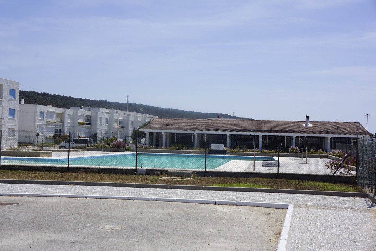 A spacious community pool is surrounded by a fence, with a large building visible on one side. The pool area includes lounge chairs and vibrant landscaping, enhancing the outdoor environment. The surrounding area features white low-rise apartment buildings and is set against a clear blue sky.