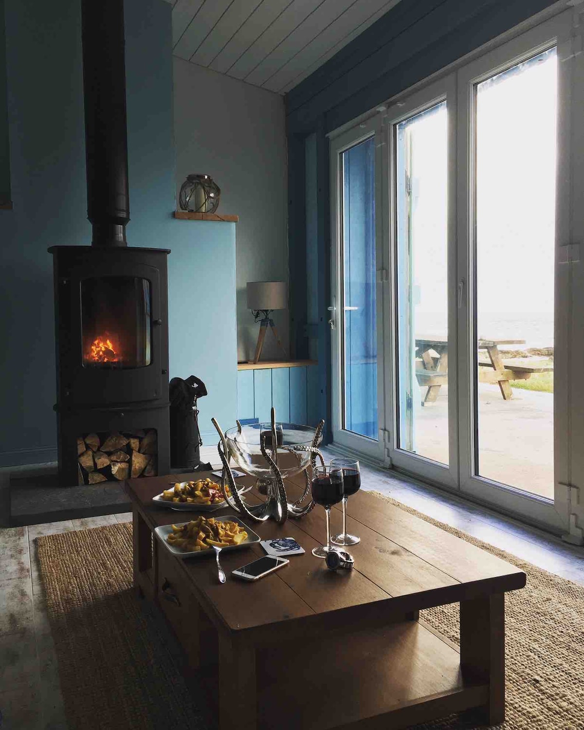 A cozy lounge area is highlighted by a log burner, providing warmth. A wooden coffee table is set with glasses and a dish of food. Large glass doors open to reveal outdoor seating with sea views, enhancing the inviting space.
