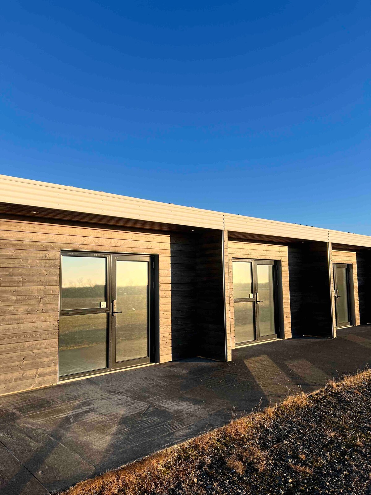 Multiple guest room entrances are lined along a wooden facade, showcasing large glass doors that reflect the sky. A smooth, dark pathway leads to each entrance, while natural surroundings are visible in the background under a clear blue sky.
