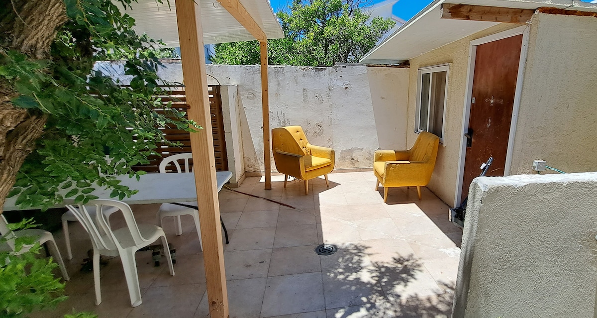 A private outdoor terrace is presented with a tiled floor and a wooden pergola for shade. Two inviting yellow armchairs are positioned around a small table, while a white dining table with plastic chairs is also visible. Surrounding walls are minimally adorned.