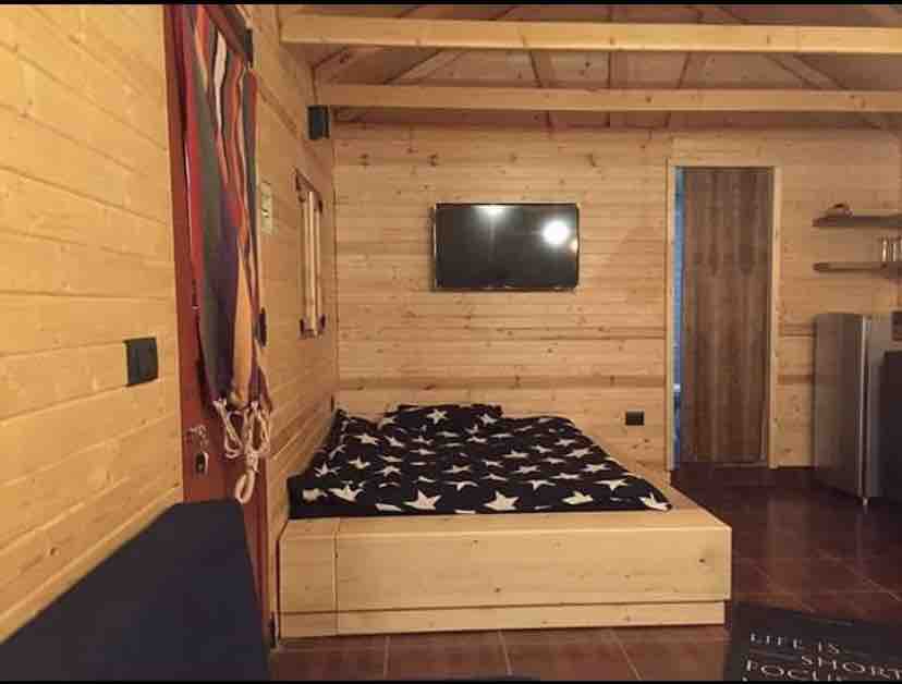 A wooden interior features a spacious bed with a star-patterned blanket. A flat-screen television is mounted on the wall, providing entertainment. The warm tones of the wood walls contribute to a cozy atmosphere, with a door leading to another area visible in the background.