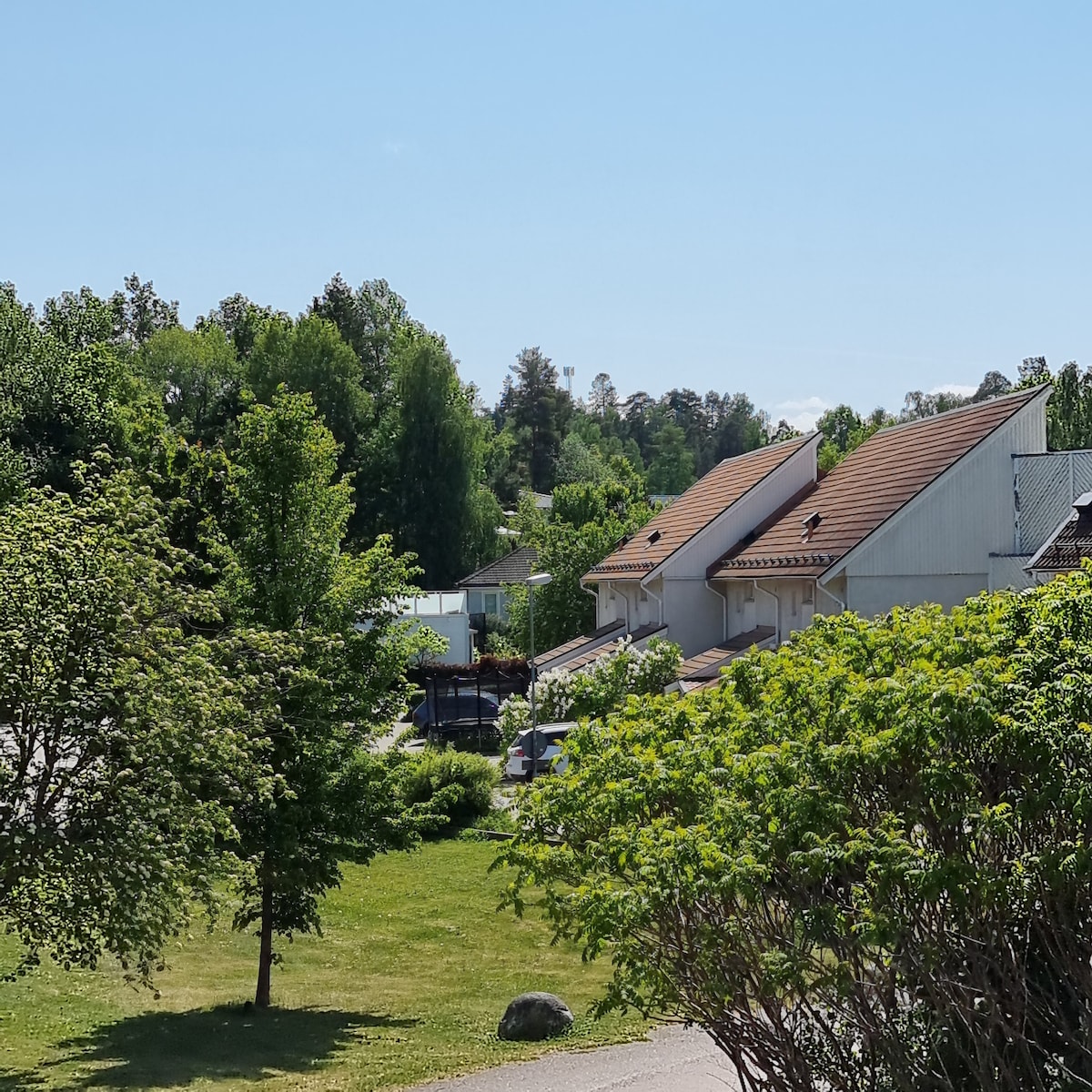 The image captures a serene residential area, featuring several houses nestled among lush greenery. Trees and shrubs provide a natural backdrop, while bright blue skies enhance the peaceful atmosphere. The arrangement of homes suggests a community setting with well-maintained landscapes.