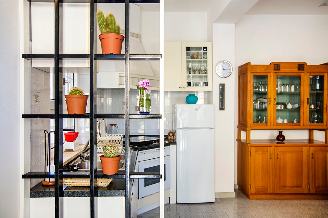 The kitchen area features a modern design with potted plants atop a divider, offering a glimpse of the cooking space. Adjacent, a wooden cabinet holds glassware and kitchen items, complementing the bright and functional environment.