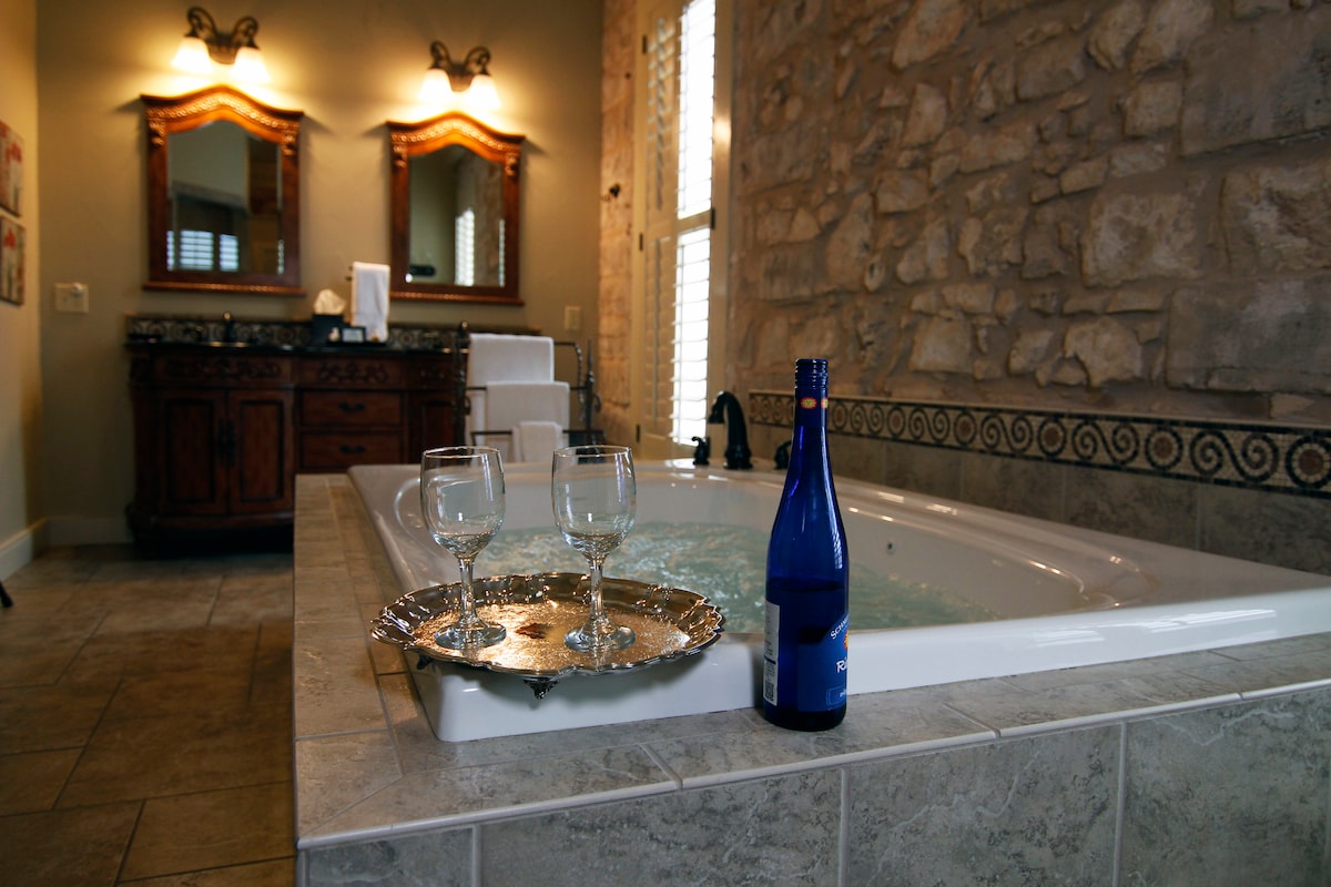 A spacious bathroom features a large, jetted tub surrounded by elegant tile work. A bottle of sparkling water sits on a silver tray alongside two glasses. The room is enhanced by natural light streaming in from the window, and the wall showcases beautiful exposed stone.