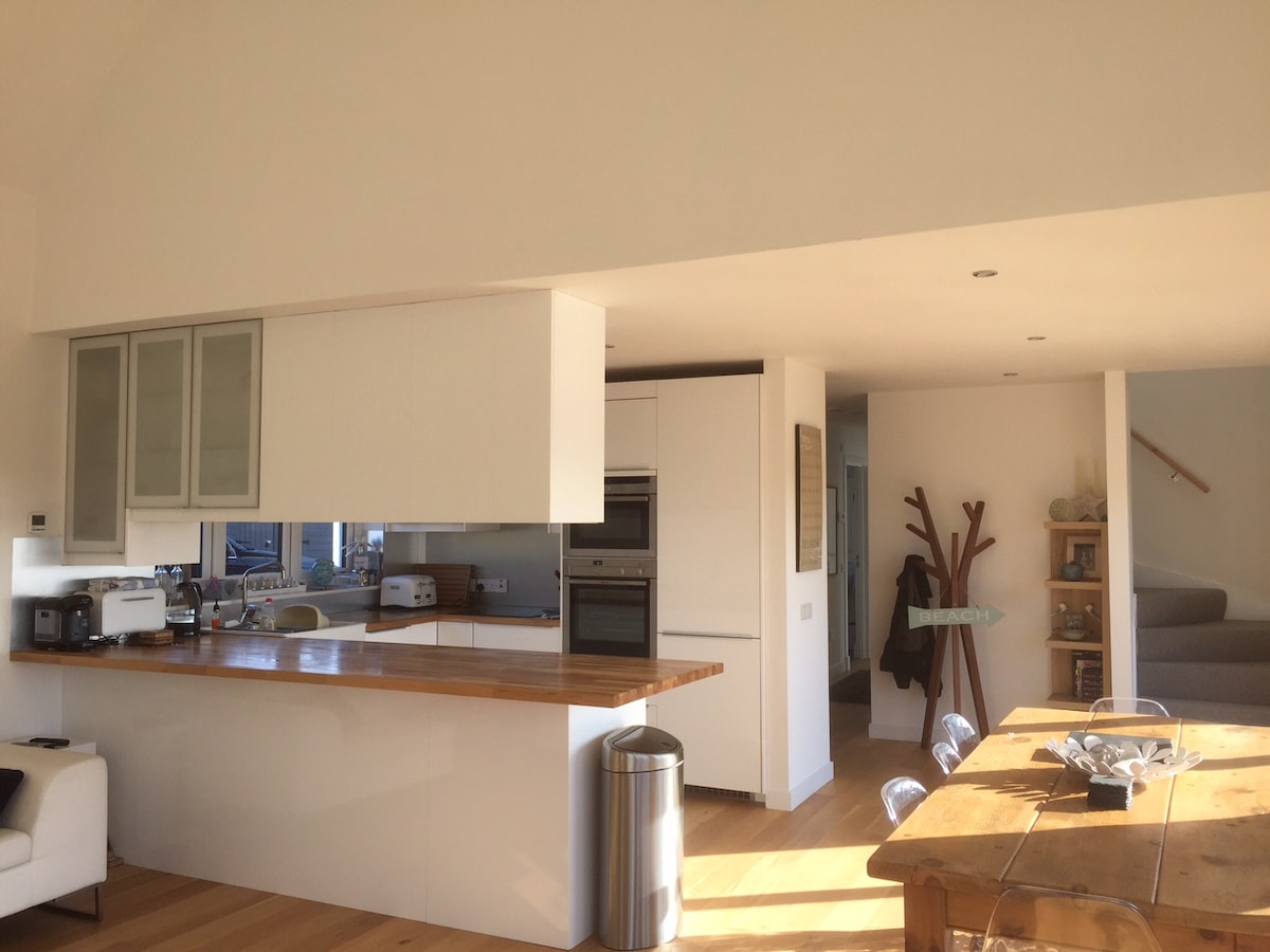The open-plan kitchen and dining area feature modern cabinetry and a spacious wooden countertop. A dining table is situated nearby with light streaming in from large windows, illuminating the wooden flooring. Minimalist decor adds to the contemporary feel of the space, creating a seamless flow to the living area.