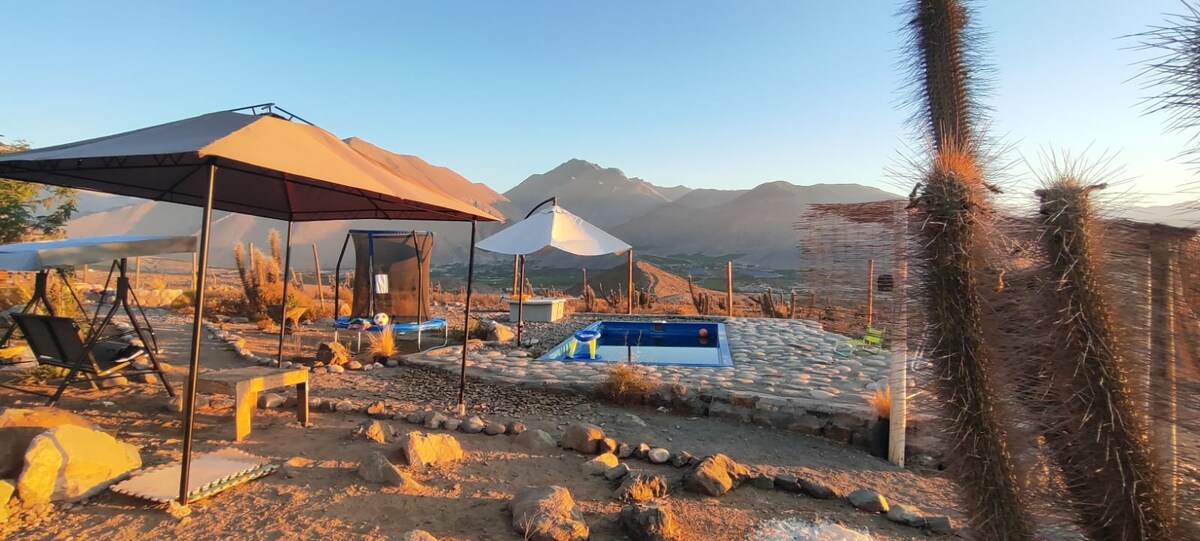 A serene outdoor area features several shaded structures flanking a small pool. The landscape is dominated by mountains in the background, illuminated by warm sunset tones. Stone pathways and natural desert flora enhance the tranquil setting, providing a welcoming environment for relaxation.