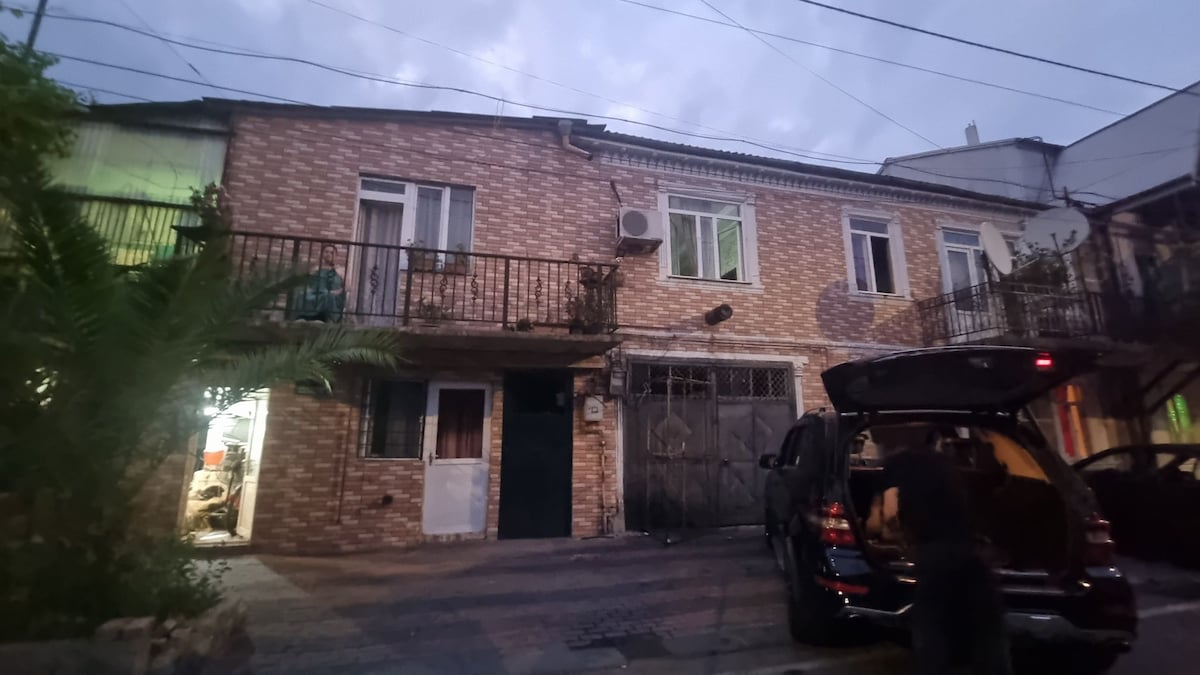 An exterior view of a two-story brick building is shown, with several windows and balconies visible. A vehicle is parked in front, while the entrance is marked by a door with a decorative gate. Surrounding greenery adds a touch of nature to the scene.