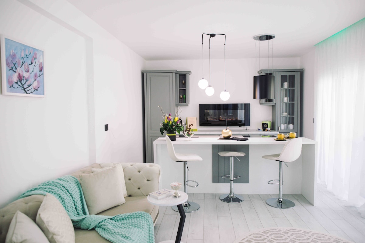 A modern kitchen and living area is featured, showcasing a light-colored sofa adorned with a soft throw. Three elegant bar stools are positioned at a white kitchen island, complemented by a floral centerpiece and various kitchen appliances. Natural light filters through sheer curtains.