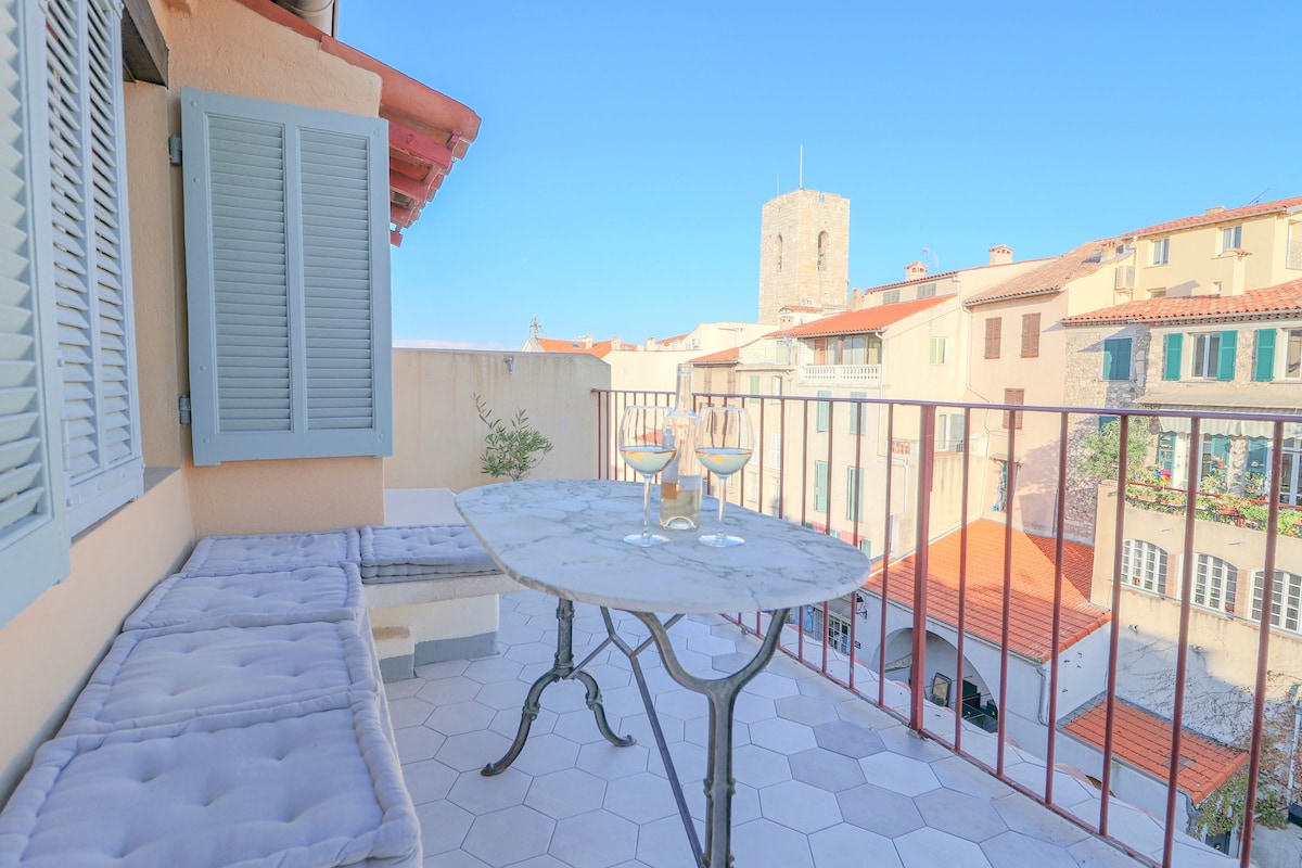 A charming terrace is presented, featuring a round marble table adorned with two glasses. Scenic views of the surrounding rooftops and a historical tower are visible in the background, framed by soft blue shutters on the left.