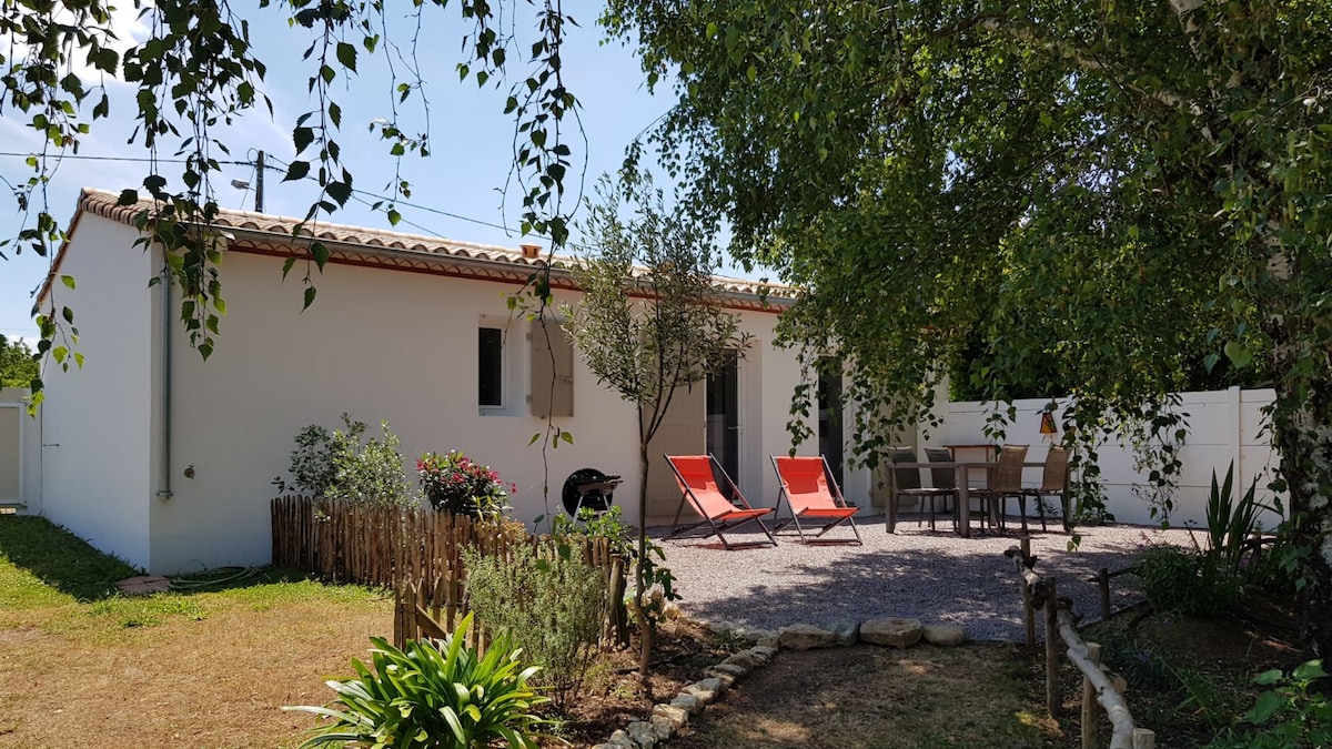 An outdoor area features a cozy seating arrangement with two bright red lounge chairs and a dining table surrounded by greenery. The charming facade of the house is visible, along with a gravel pathway leading to the entrance. A tree provides shade to the space.